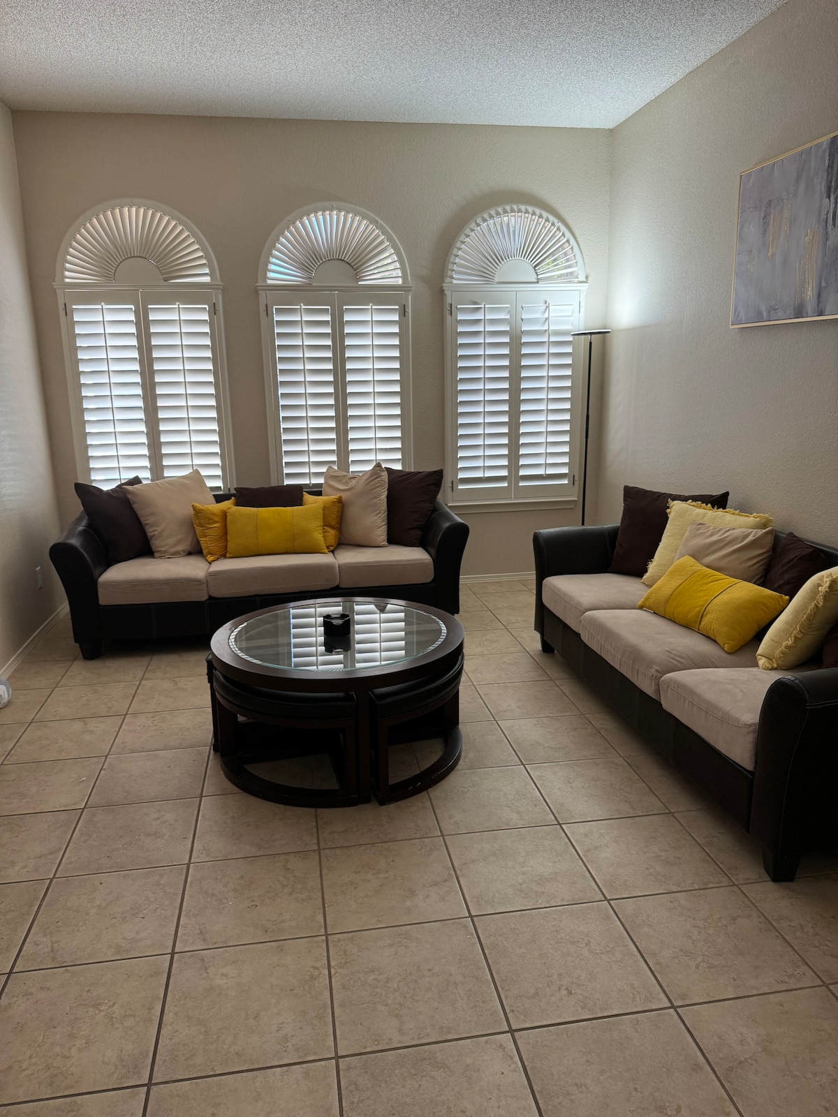 A bright living room features two comfortable couches adorned with yellow accent pillows. A round coffee table sits in the center, and light filters through elegant shuttered windows, creating a warm ambiance in the space.