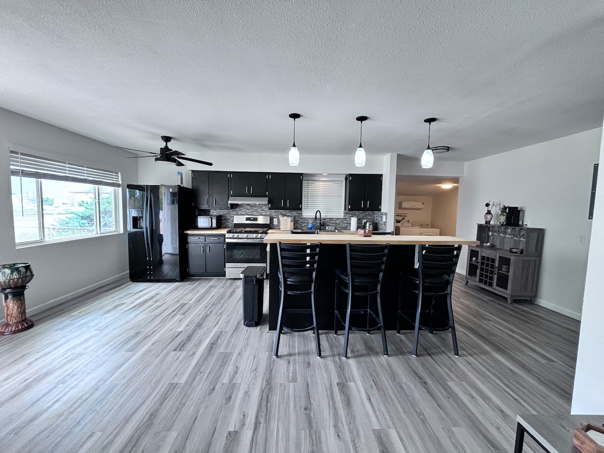 A spacious kitchen opens to a dining area, featuring a large island with seating for several guests. Dark cabinetry contrasts with light countertops. Large windows provide natural light, and sleek appliances are arranged for easy access, enhancing the functionality of the space.