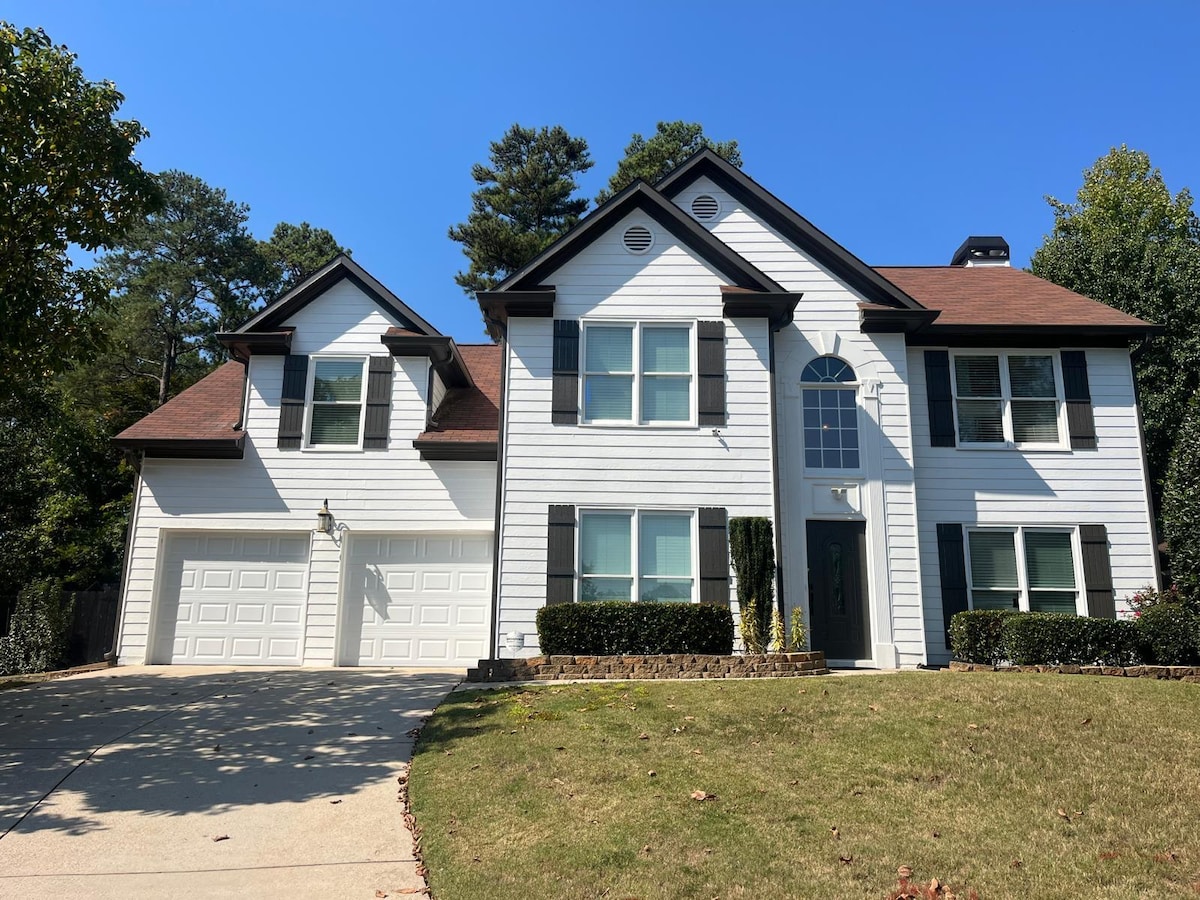 The exterior of a spacious two-story house is displayed, featuring a combination of white siding and dark shutters. A two-car garage is visible, along with a well-maintained lawn and neatly trimmed bushes. Clear blue skies provide a bright backdrop to the scene.