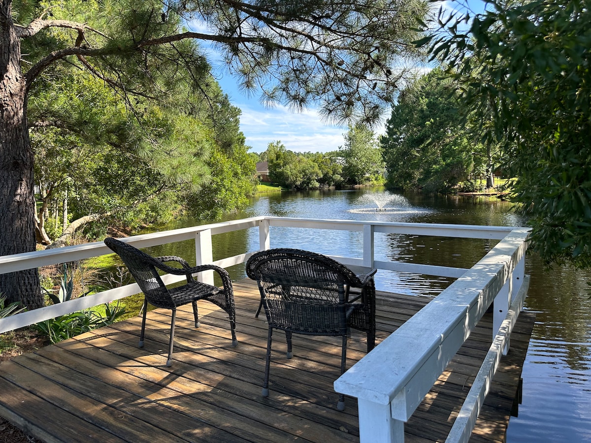A private dock extends over the serene pond, featuring two wicker chairs positioned on a wooden deck. Lush greenery surrounds the water, with a soft fountain visible in the distance under a clear blue sky.