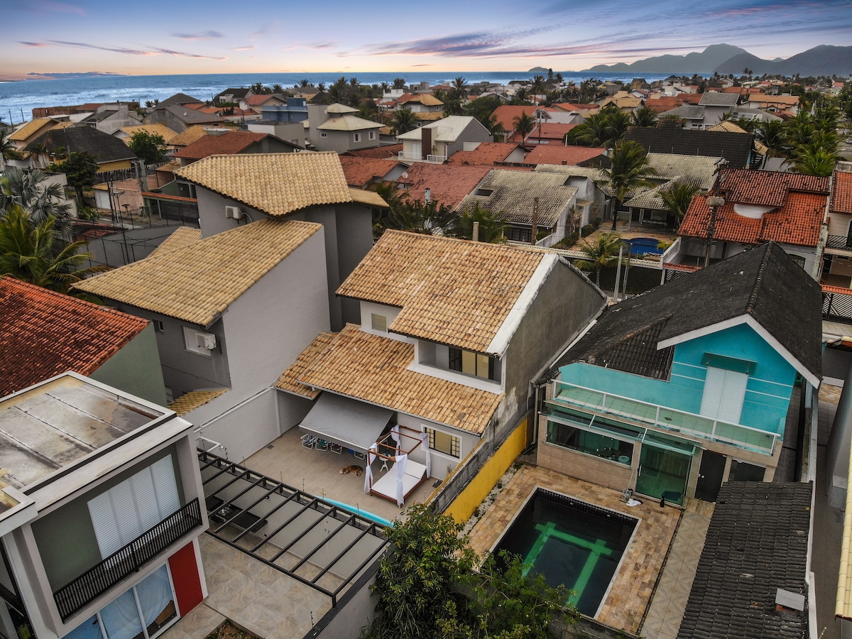 An aerial view captures a vibrant neighborhood with a variety of houses, highlighting a spacious home with a pool and outdoor area. The ocean is faintly visible in the background, suggesting proximity to the beach. A mix of rooftops adds to the charm of the setting.