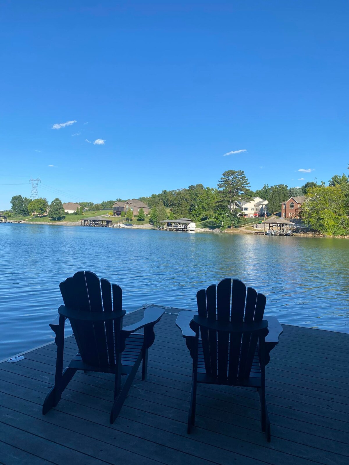Two dark Adirondack chairs are positioned on a wooden dock, overlooking a calm lake. The water reflects a clear blue sky with scattered clouds. Lush greenery and residential homes border the opposite shore, creating a serene lakeside setting.