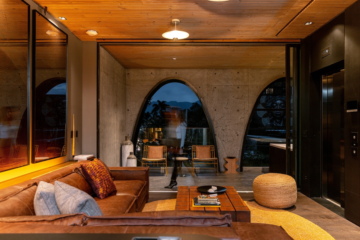 A modern living space features a cozy brown sofa with decorative pillows, paired with a wooden coffee table. Large arched windows provide views of the outdoor landscape. Natural materials and a warm ceiling light create a welcoming ambiance, complemented by stylish seating in the background.