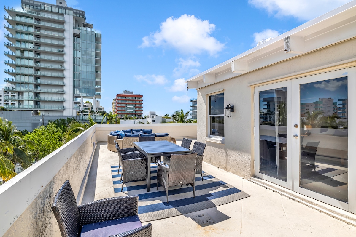 A spacious outdoor terrace is depicted, featuring a large dining table surrounded by comfortable wicker chairs. The area is adorned with a soft rug, providing a welcoming touch. A panoramic view of Miami's skyline is visible in the background, enhancing the coastal atmosphere.