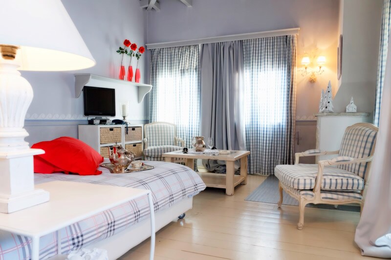 A small suite features pastel grey-blue walls, with a cozy seating area furnished by a striped chair and a small table. Natural light floods through two curtained windows, and decorative elements include a lamp and small vases with red roses. A comfortable bed is visible in the foreground.