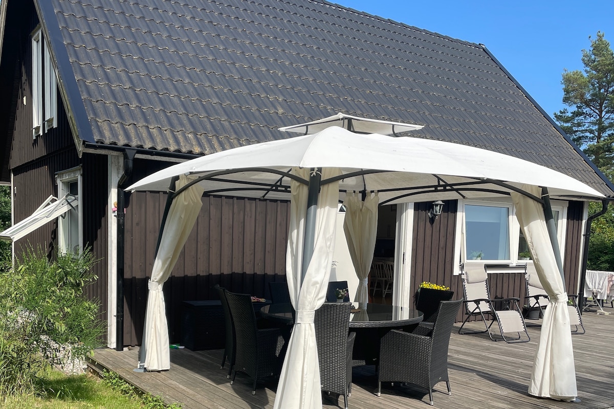 A covered outdoor seating area is featured, surrounded by natural greenery. Woven chairs and a circular table are present under the canopy. The clear sky reflects on the textured roof of the house, which stands to the side. Sunlight enhances the inviting space.