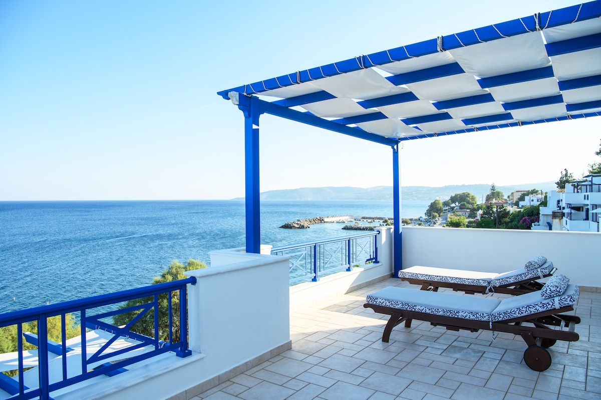 A spacious terrace is visible, furnished with sun loungers featuring patterned cushions. A large, shaded area provides comfort, overlooking the tranquil blue sea and distant coastline, enhancing relaxation. The vivid blue framework complements the serene environment, inviting guests to enjoy the outdoor space.