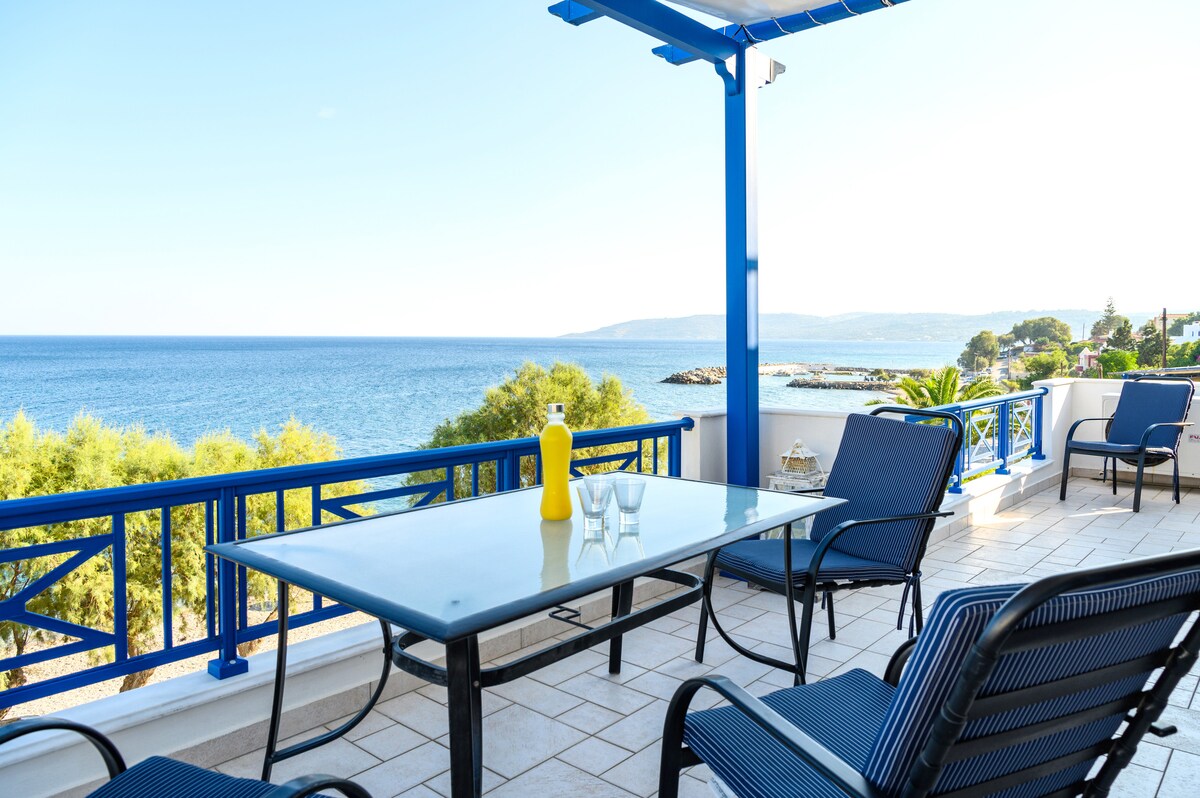 A spacious balcony is highlighted, featuring a glass dining table surrounded by comfortable chairs. A vibrant blue railing frames the view of the serene sea and distant coastline, creating an inviting outdoor space for relaxation and meals.