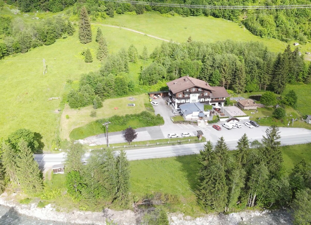 Aerial view of a large building complex surrounded by lush greenery. The structure features multiple levels and is complemented by a parking area. A road runs alongside the property, bordered by trees and natural landscapes, offering a serene setting.