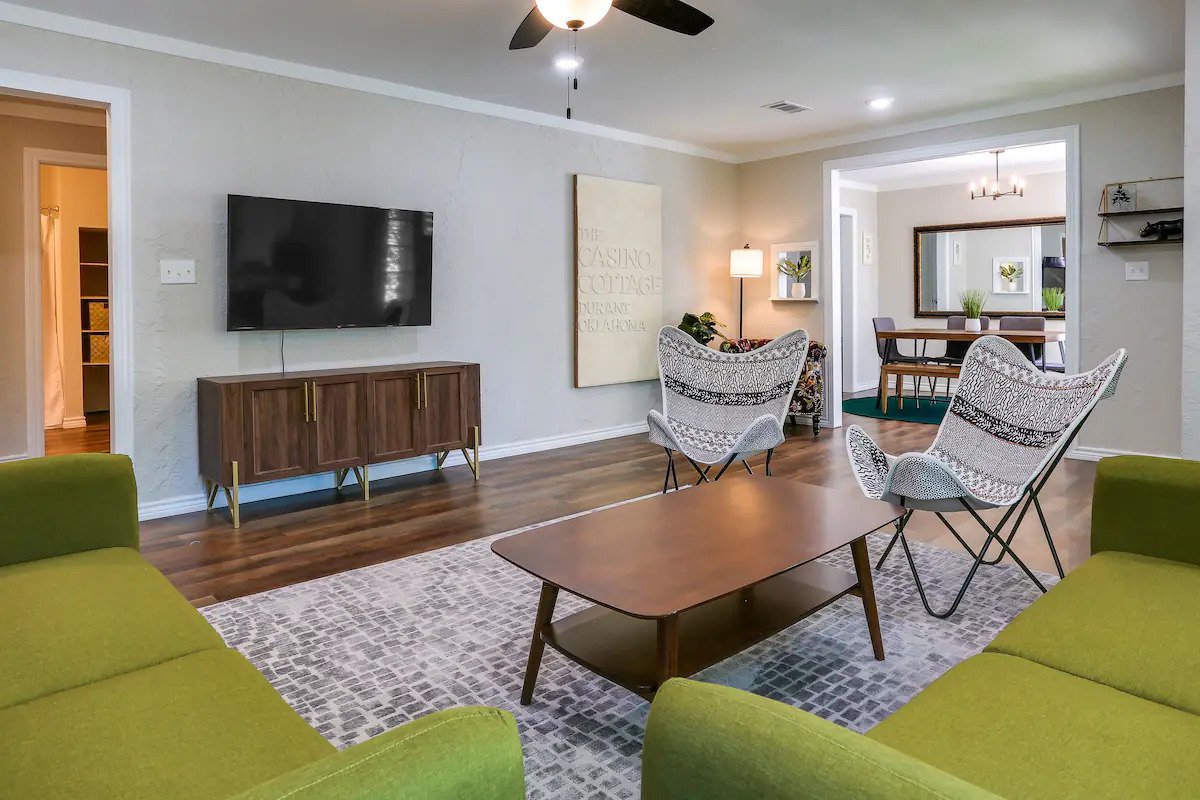 The living room presents an open layout featuring two cozy couches paired with two accent chairs. A medium-sized coffee table sits in the center, while a wall-mounted TV is positioned across from the seating. Natural light fills the space, complemented by neutral wall colors.