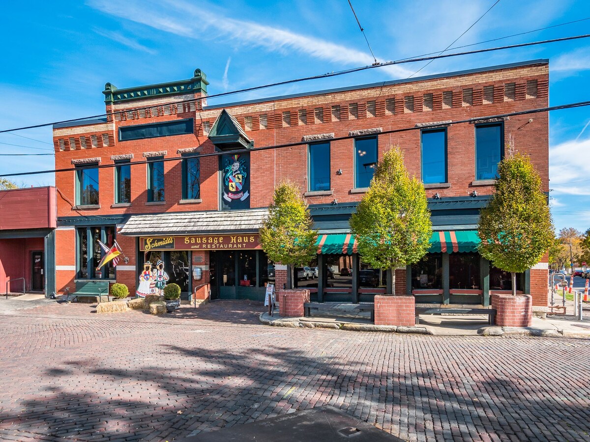 A historic brick building stands prominently, featuring large windows and an inviting entrance. Green canopies adorn the exterior, framing the restaurant's name. Paved streets lead up to the establishment, surrounded by neatly trimmed trees, creating a welcoming atmosphere.