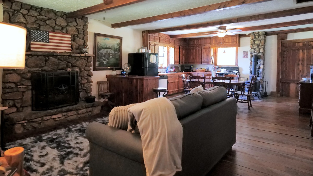 A spacious living area features rustic stone walls and wooden beams. A comfortable couch sits atop a soft area rug, with a stone fireplace as a focal point. The open layout includes an adjacent kitchen with wooden cabinets and a dining area with multiple chairs.