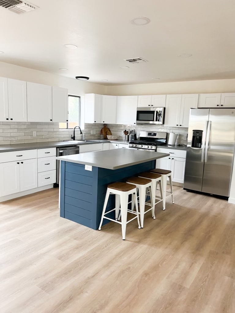 A newly remodeled kitchen features white cabinetry paired with a stylish blue island. Stainless steel appliances include a refrigerator and a microwave. Three bar stools are positioned at the island. Natural light streams in through the window, illuminating the light wood flooring.