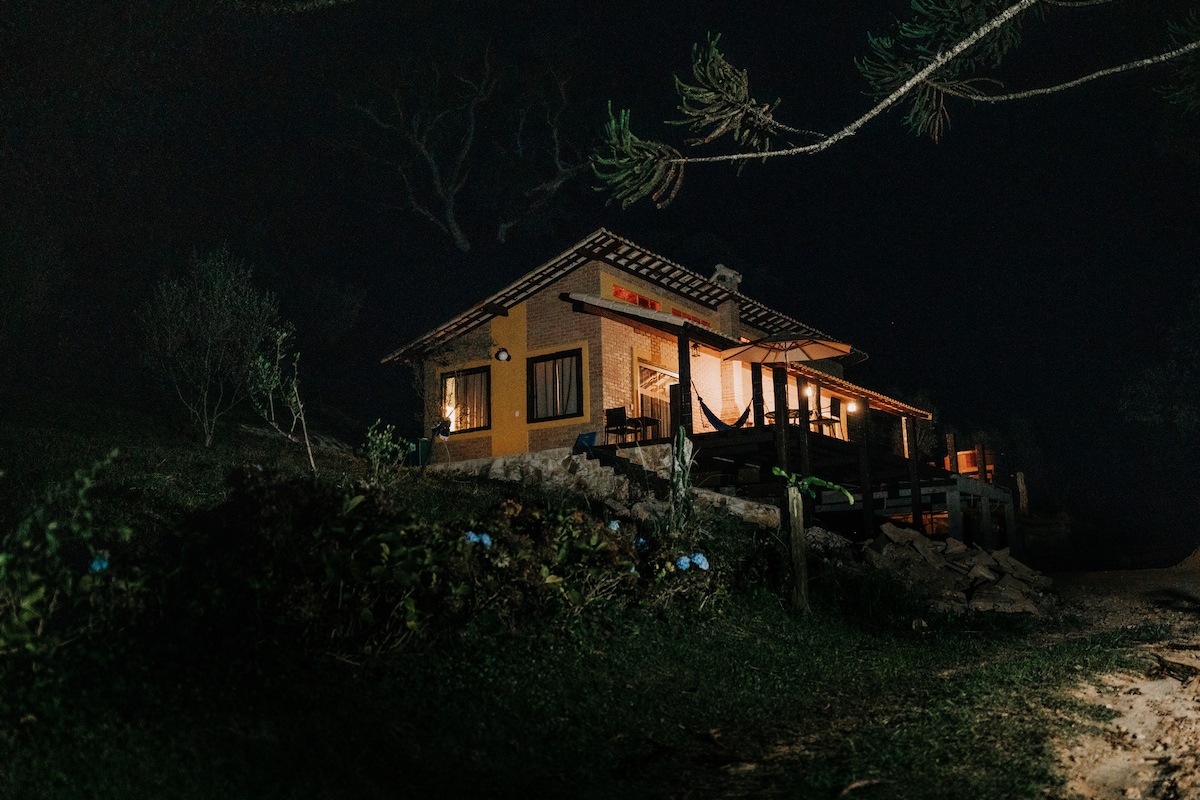 A charming chalet is illuminated at night, showcasing warm light emanating from the windows. The building features a traditional roof and is surrounded by greenery, with a pathway leading to its entrance. The tranquility of the surroundings is highlighted by the soft glow.
