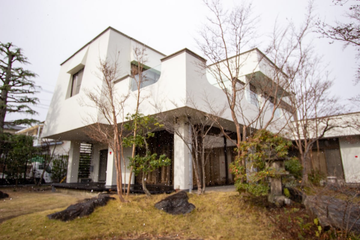 The exterior of the renovated single-family house is showcased, featuring a modern architectural design with large windows. Surrounding greenery includes small trees and a stone feature, complemented by a landscaped yard with decorative rocks.