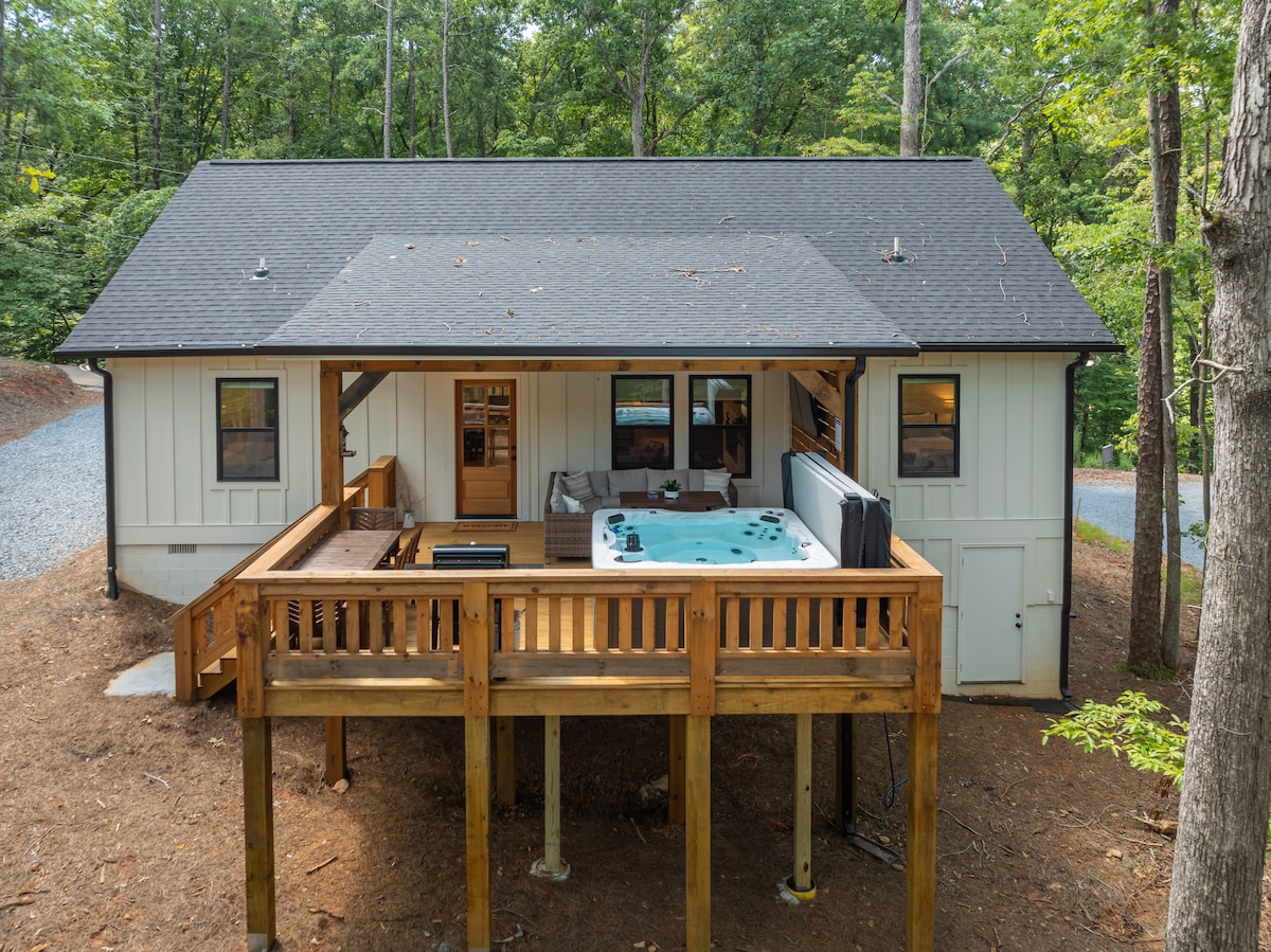 The exterior of the cottage features a spacious back deck, which includes an oversized hot tub and ample seating. The surrounding forest provides a natural backdrop, while the light-colored siding and dark roofing create a modern contrast.