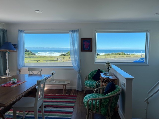 A bright living space features large windows framing scenic ocean views. A wooden dining table is set alongside colorful chairs, with vibrant decor enhancing the ambiance. Light curtains flutter gently, inviting natural light, while a striped rug adds warmth to the room.