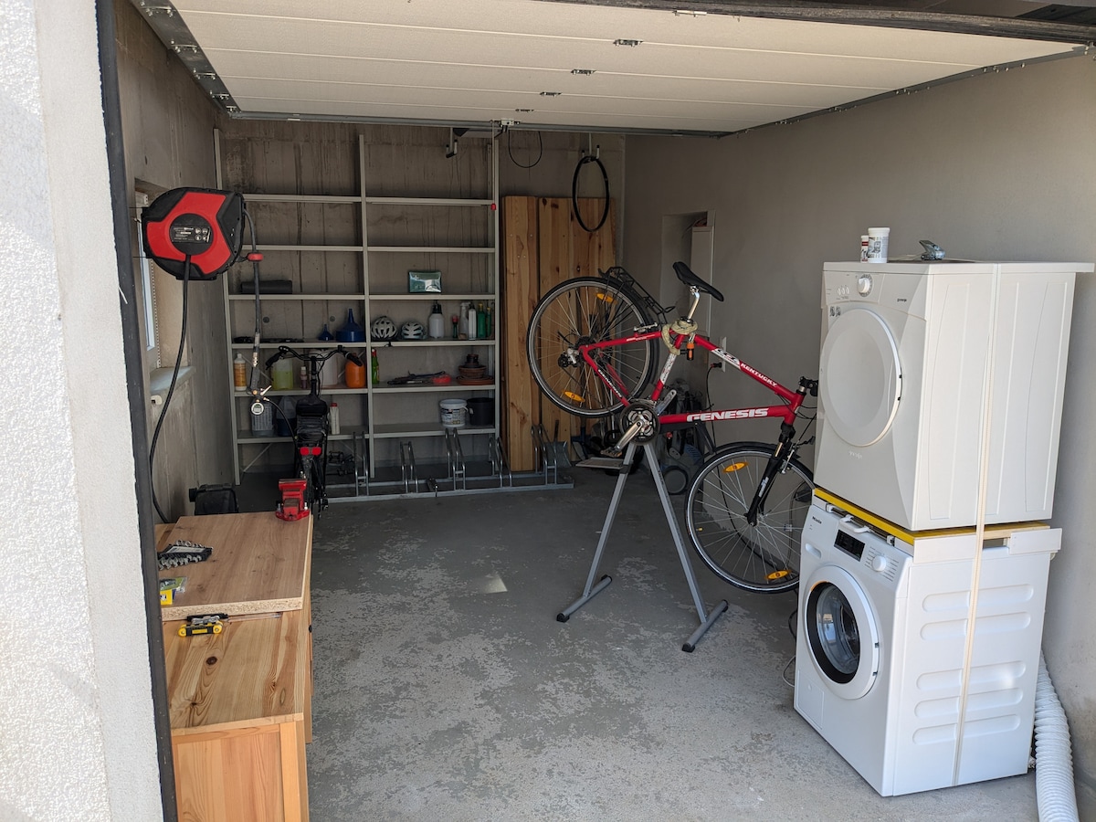 A functional garage space is presented, featuring a bicycle mounted on a stand and a washing machine. A wooden workbench sits against one wall, and a shelving unit displays various tools and equipment. The floor is unpolished cement, contributing to a utilitarian environment.