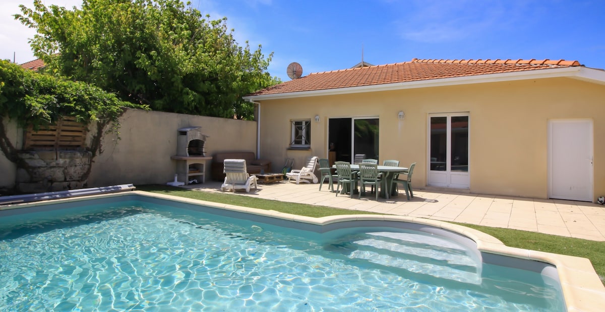 A spacious outdoor area features a clear pool surrounded by tiled flooring. A table with chairs is positioned nearby, and two lounge chairs are arranged next to the pool. The house exterior showcases large windows allowing light to enter, complemented by a shaded green area.