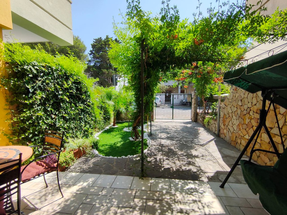 The outdoor area features a stone-paved patio with a small round table and two black chairs. A swing is positioned to the right under a green canopy. Lush greenery surrounds the space, and a gated entrance provides access to the street.