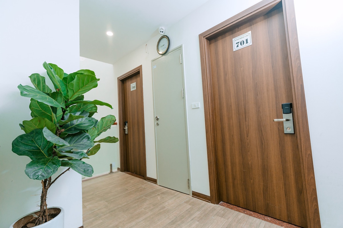 A well-lit hallway features contemporary wooden doors, each marked with room numbers. A decorative potted plant adds a touch of greenery, enhancing the inviting atmosphere. The flooring is light-colored, and a wall clock is mounted above one of the doors, contributing to the functional design.