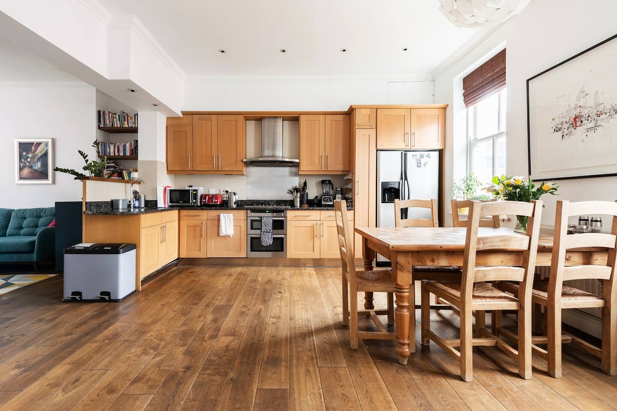 A modern kitchen and dining area showcases wooden cabinetry and a large table surrounded by six wooden chairs. Stainless steel appliances, including a refrigerator and oven, are present. Natural light pours in through a window, highlighting the spacious, open layout and wooden flooring.