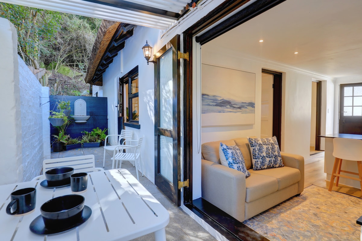 A cozy living area featuring a comfortable sofa adorned with decorative pillows. Large glass doors open to an inviting outdoor space, while a dining table is set with black plates and mugs. Natural light brightens the interior, highlighting the light-colored decor.