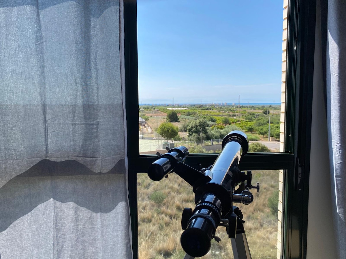 A telescope positioned beside a window captures a view of lush greenery and distant landscapes. Natural light filters through sheer curtains, illuminating the inviting space. The horizon extends into the background, indicating a peaceful environment.