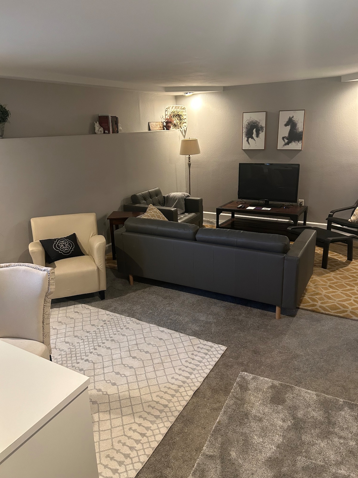 A cozy living area features a gray sectional sofa and two armchairs, arranged around a coffee table. A flat-screen television is positioned on a wooden stand against the wall. Soft lighting enhances the neutral color scheme, while a patterned area rug adds texture to the space.