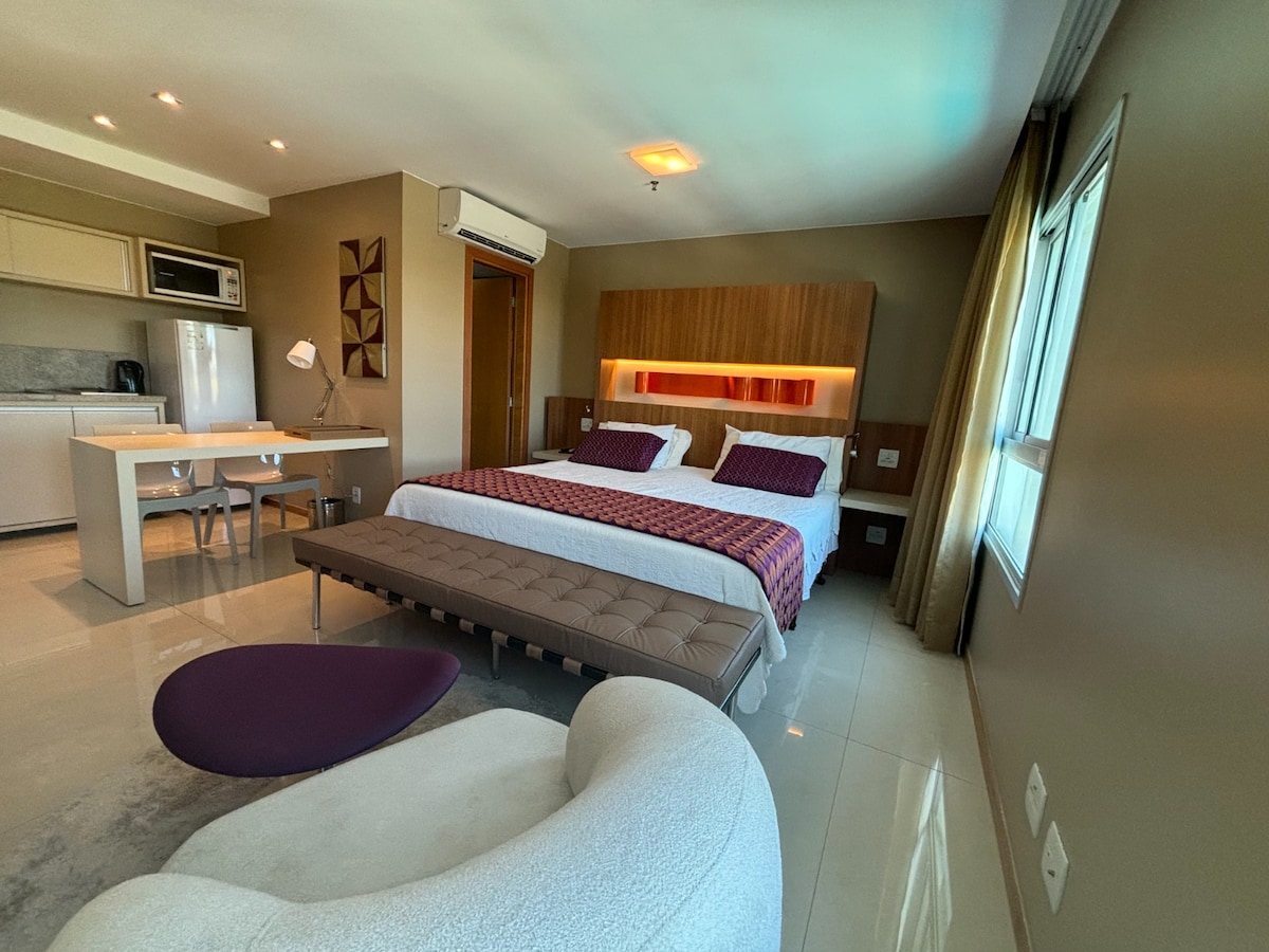 The room features a spacious bed with decorative pillows, complemented by a comfortable lounge chair and contemporary table. A kitchenette area with appliances sits to the left, while natural light filters through the window, enhancing the neutral color scheme.