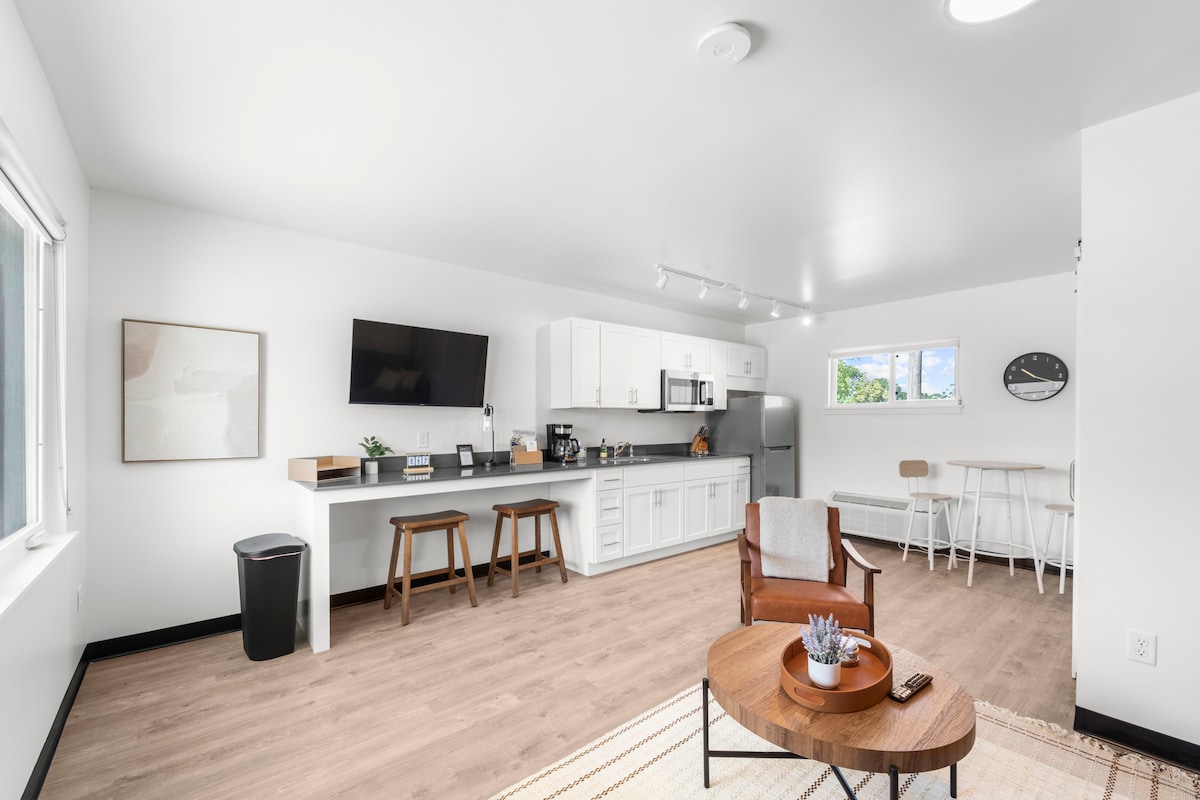 A modern open layout features a fully equipped kitchen with white cabinetry and stainless steel appliances. A television is mounted on the wall, and a mix of seating options, including a wooden table and light chairs, creates a flexible living space. Large windows provide natural light.