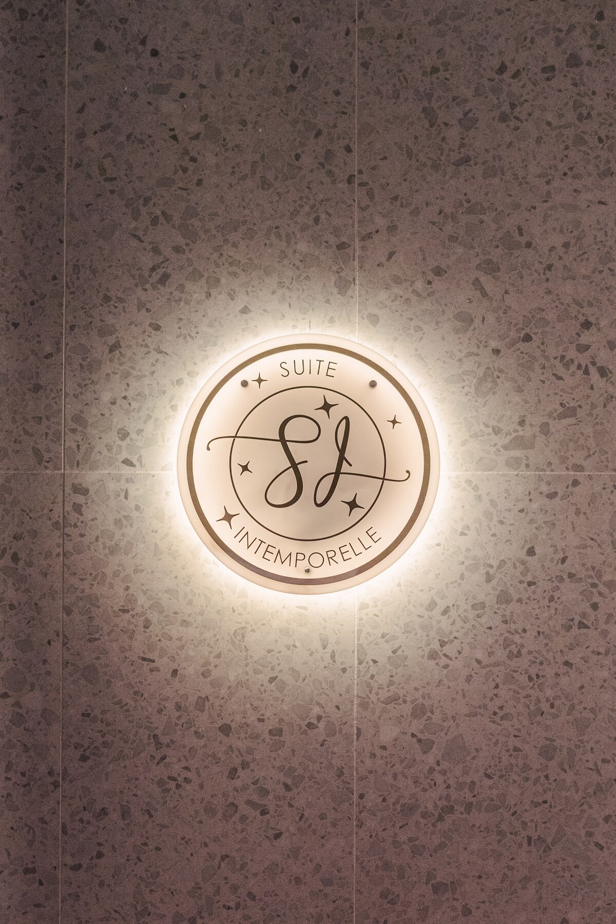 A circular wall light illuminates the logo of the 'Suite Intemporelle' against a textured, light-colored stone wall. The soft glow enhances the branding, creating an inviting entry point into the suite.