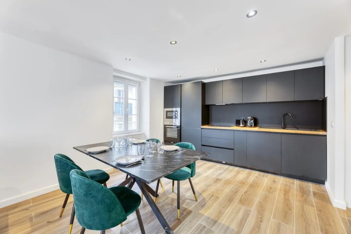 An open kitchen and dining area features a modern design with a sleek black cabinetry against light, airy walls. A dining table with four green velvet chairs occupies the center, while high-end appliances are arranged neatly on the countertop.