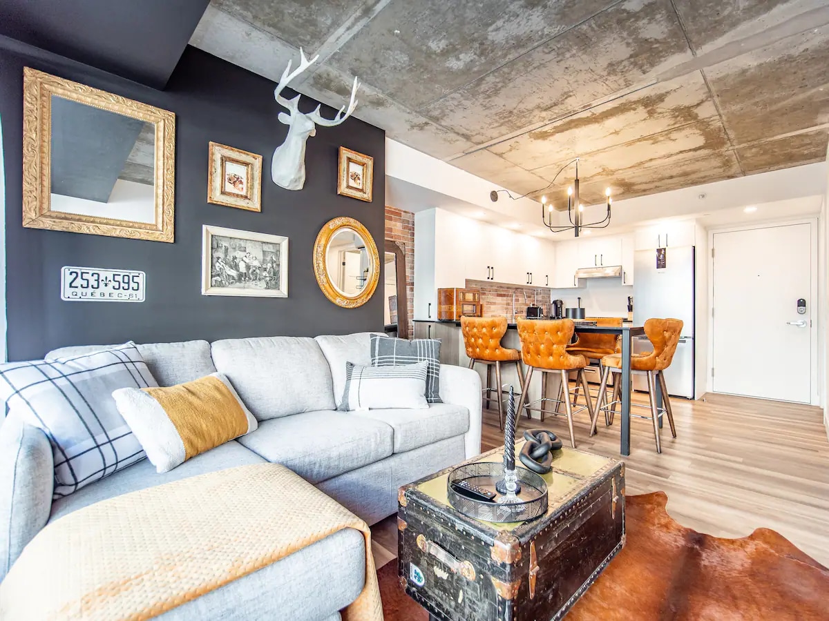 A modern living space features a gray sofa adorned with a cozy throw and a decorative cushion. Warm lighting creates an inviting atmosphere. Stylish orange chairs sit at a dining table, and artwork hangs on the textured walls, complementing the elegant decor.