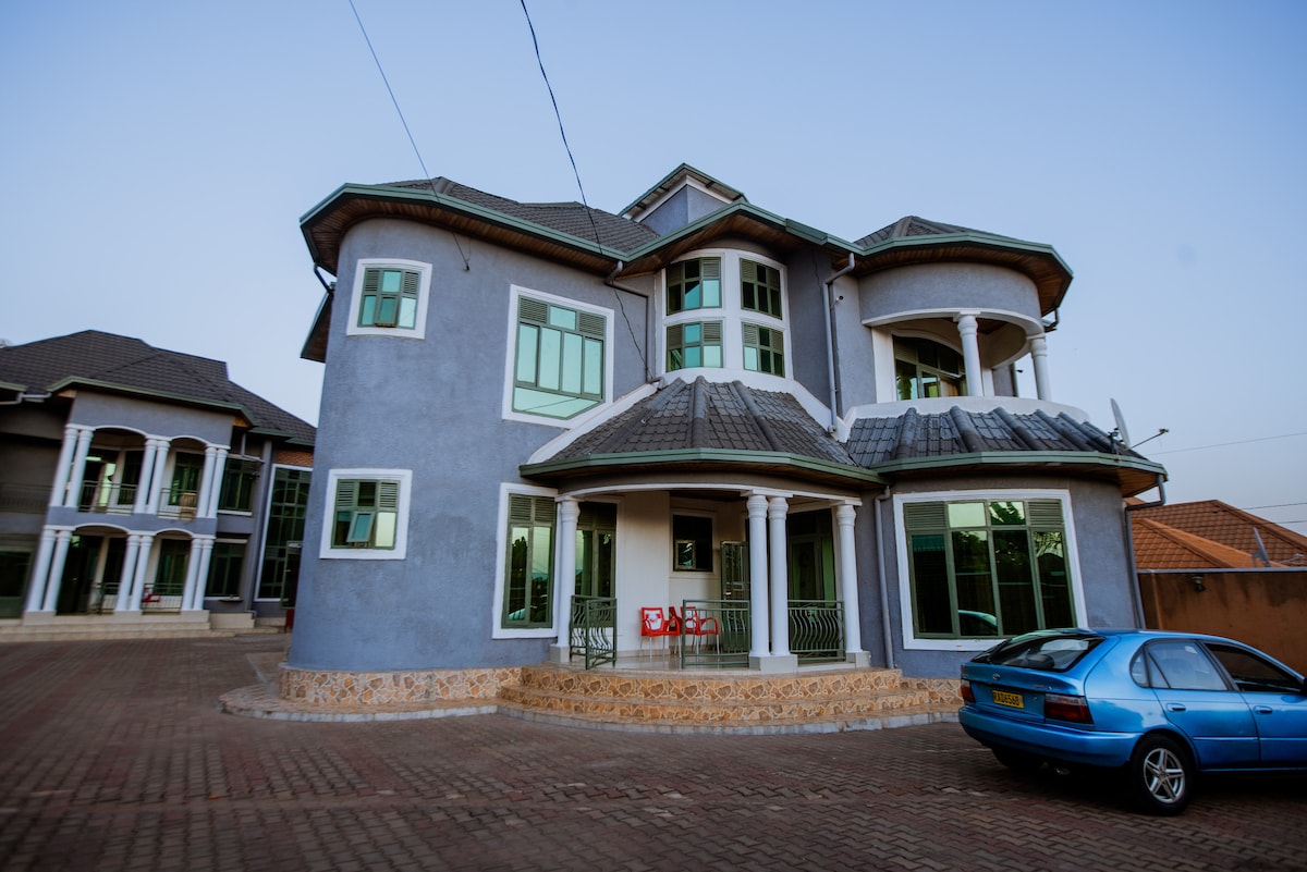A spacious two-story house features a unique architectural design with rounded corners and multiple large windows. The exterior is painted in soft blue, complemented by a decorative stone base. A parked car is visible in the driveway, enhancing the welcoming entrance.