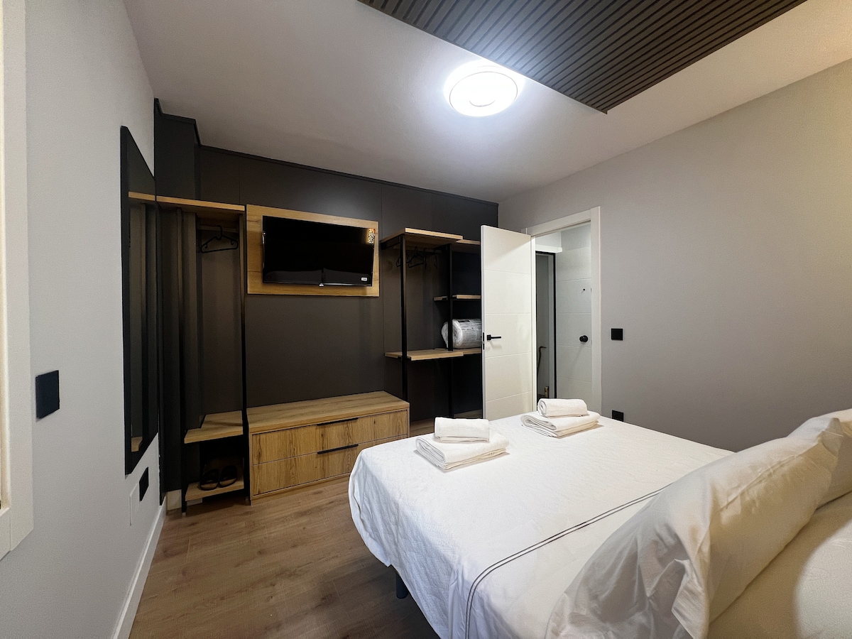 A well-organized bedroom features a spacious bed with neatly arranged linens. A wooden bench and wardrobe provide ample storage, while a wall-mounted television offers entertainment options. Soft lighting highlights the modern design elements, contributing to a calm atmosphere.