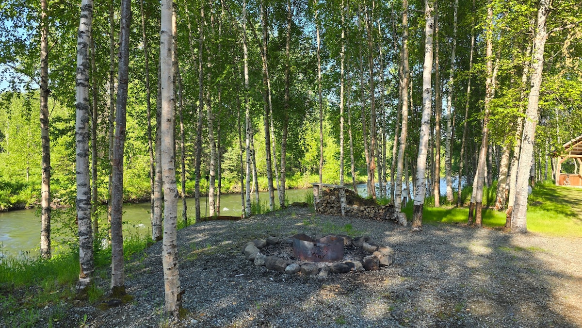 A serene campfire area is surrounded by tall trees with lush green leaves, providing shade and a natural ambiance. A stone fire pit is centered on a gravel ground, and the tranquil Little Susitna River can be seen in the background.