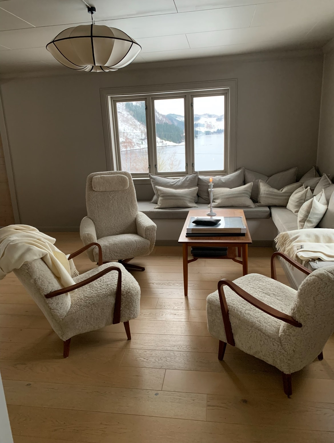 A bright sitting area features two textured armchairs and a cozy window seat filled with cushions. A small wooden table is positioned in the center, topped with a candle and books. Large windows provide a view of the scenic outdoors.