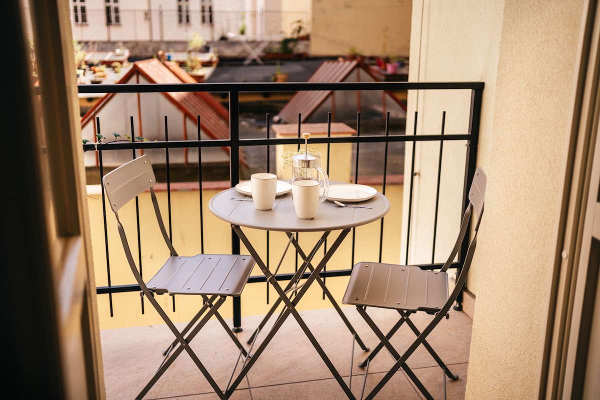 A small balcony features a round table with two folding chairs. On the table, two cups and a glass carafe are arranged. The view includes rooftops of nearby buildings, adding to the urban setting.