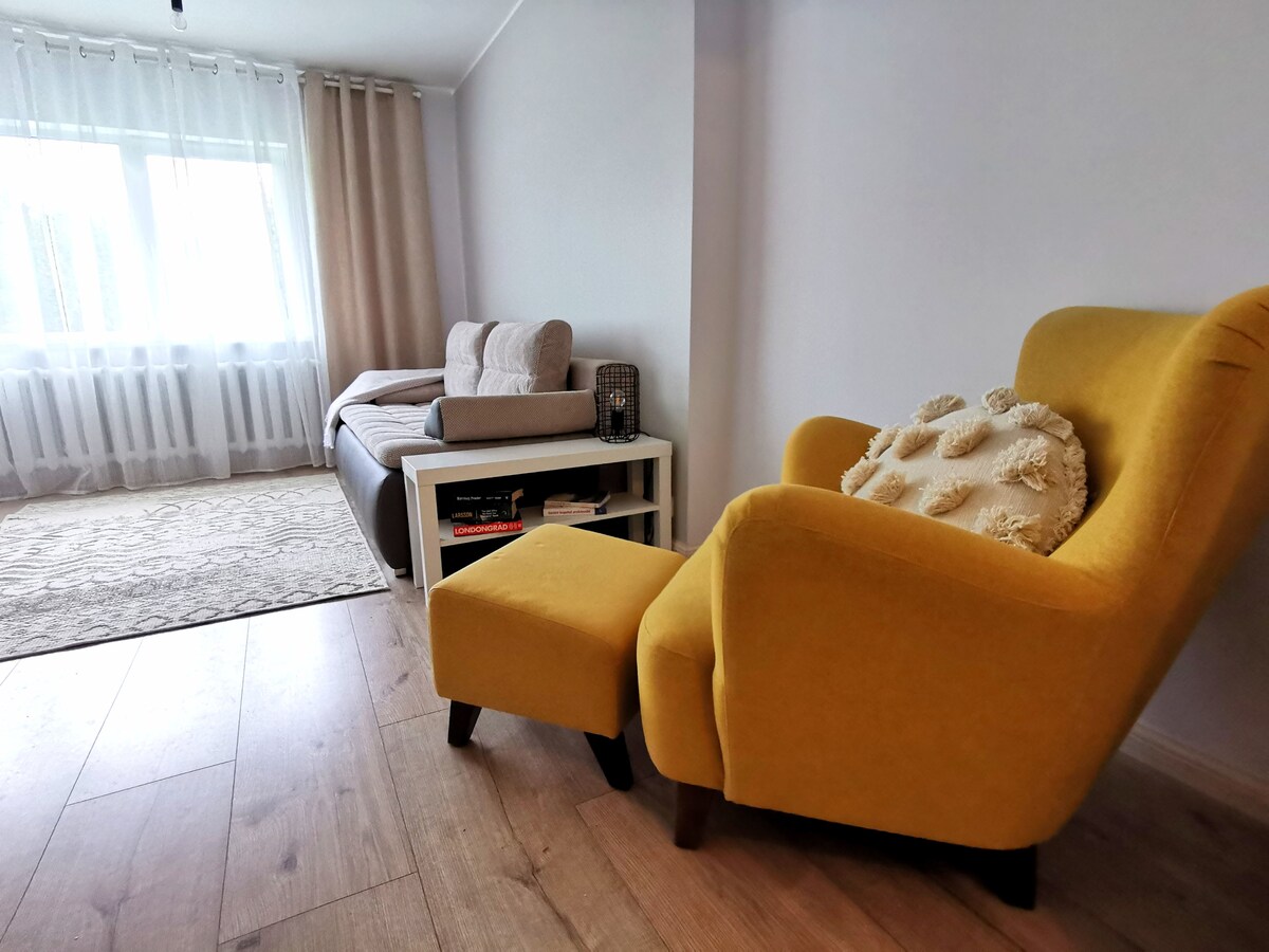 A bright and inviting living space features a comfortable yellow armchair paired with a matching ottoman. A neutral-toned sofa bed is positioned nearby, with large windows allowing natural light to illuminate the wooden floor. Soft curtains frame the window, creating a cozy atmosphere.