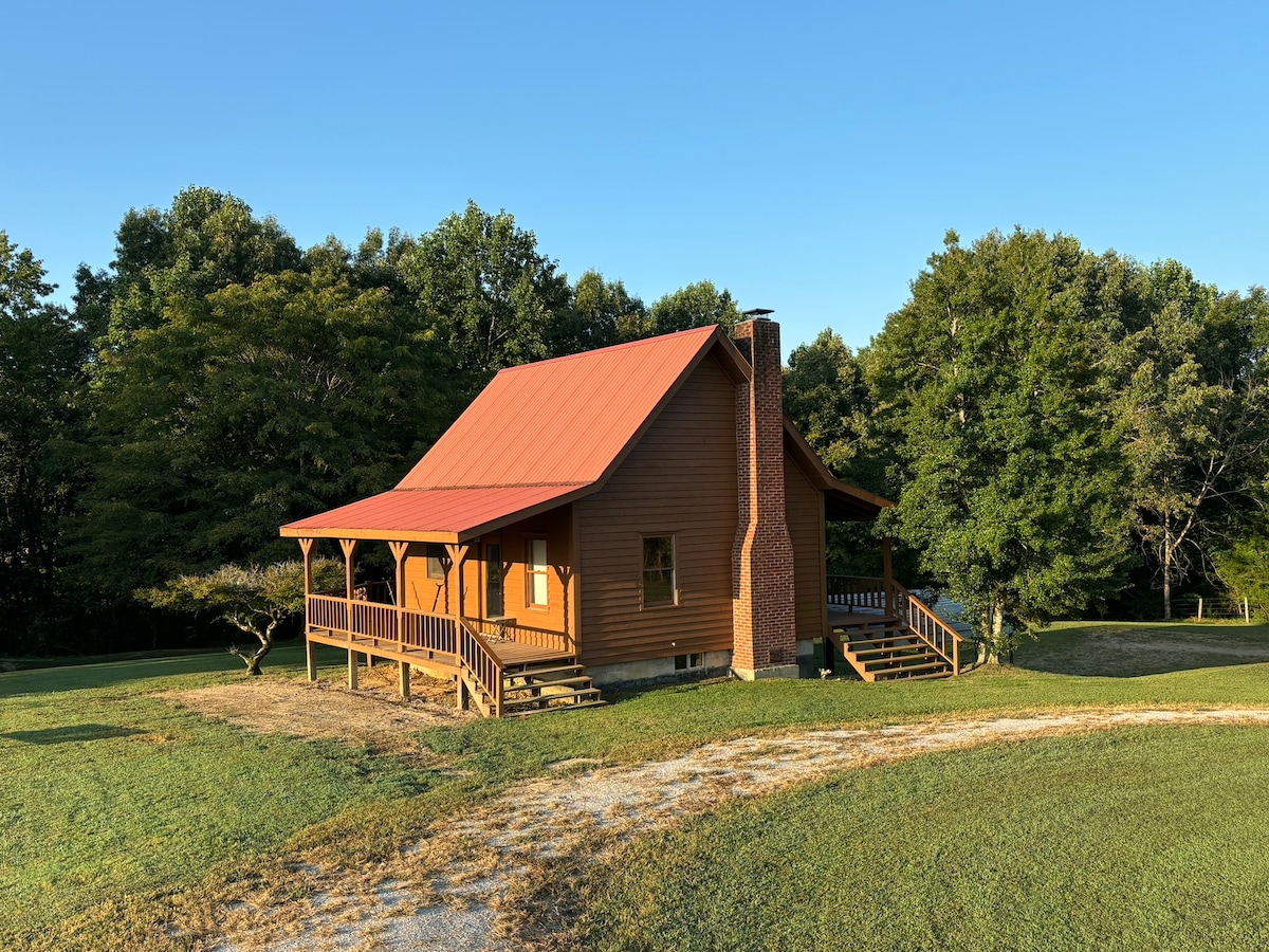 A rustic wooden cabin with a bright red roof is set amidst lush greenery. The wrap-around porch invites relaxation, while the surrounding yard offers ample outdoor space. A gravel path leads to the cabin, showcasing peaceful rural surroundings.