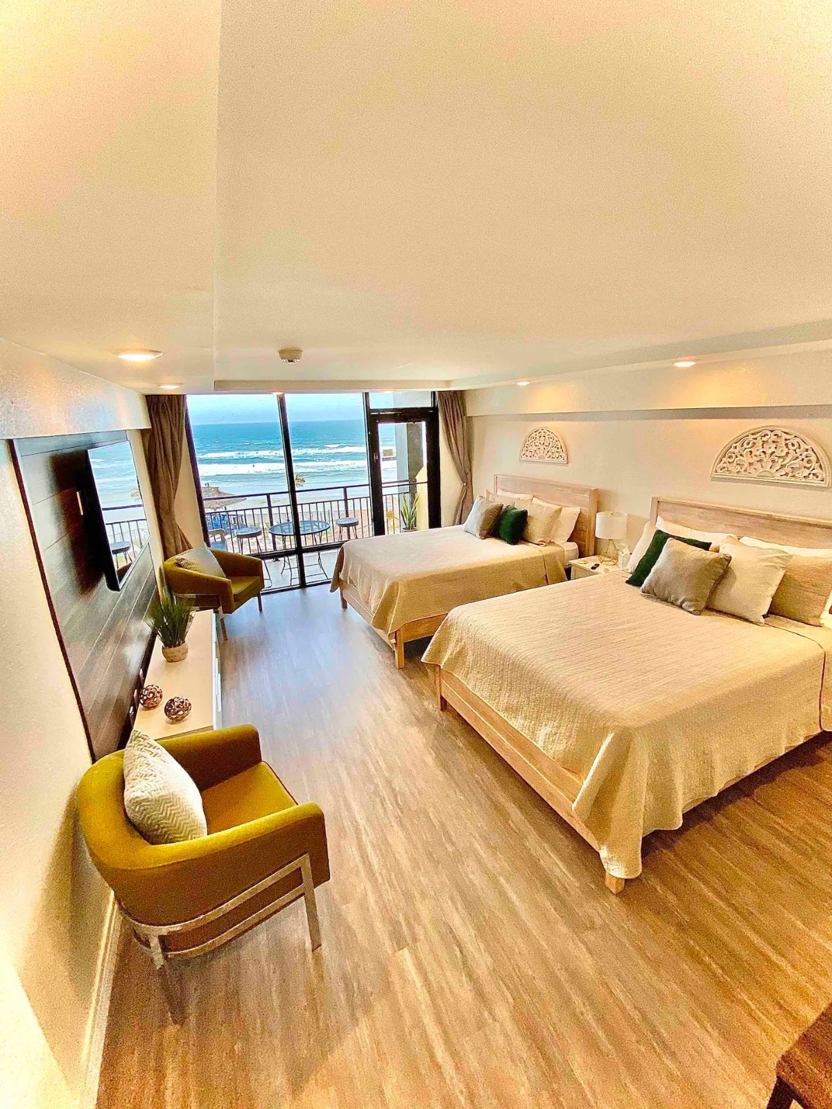 The studio features two spacious queen beds dressed in neutral linens. A large window provides ocean views, and natural light fills the room. Green accent chairs add a pop of color. A wall-mounted television and decorative elements are visible, creating a comfortable atmosphere.