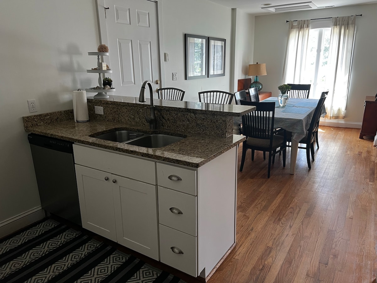 A modern kitchen area features a granite countertop with a sink, dishwasher, and ample space for meal preparation. A dining area with a large table and elegant chairs is illuminated by natural light from windows, creating a warm and welcoming environment.