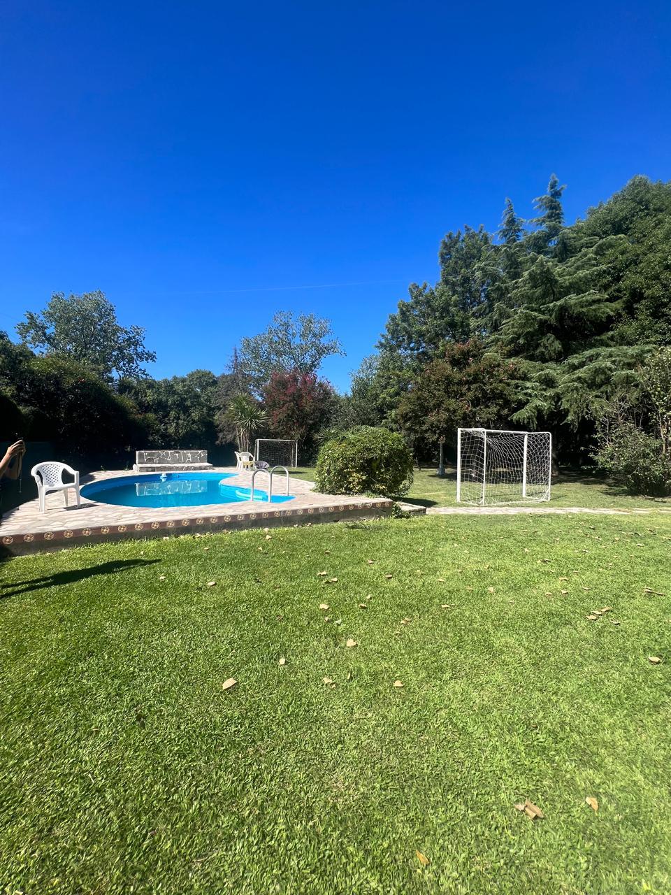 A clear blue sky stretches overhead, highlighting a spacious green lawn. A swimming pool is shown beside a stone patio, and a small goalpost is visible in the background, offering a playful addition to the serene outdoor space.