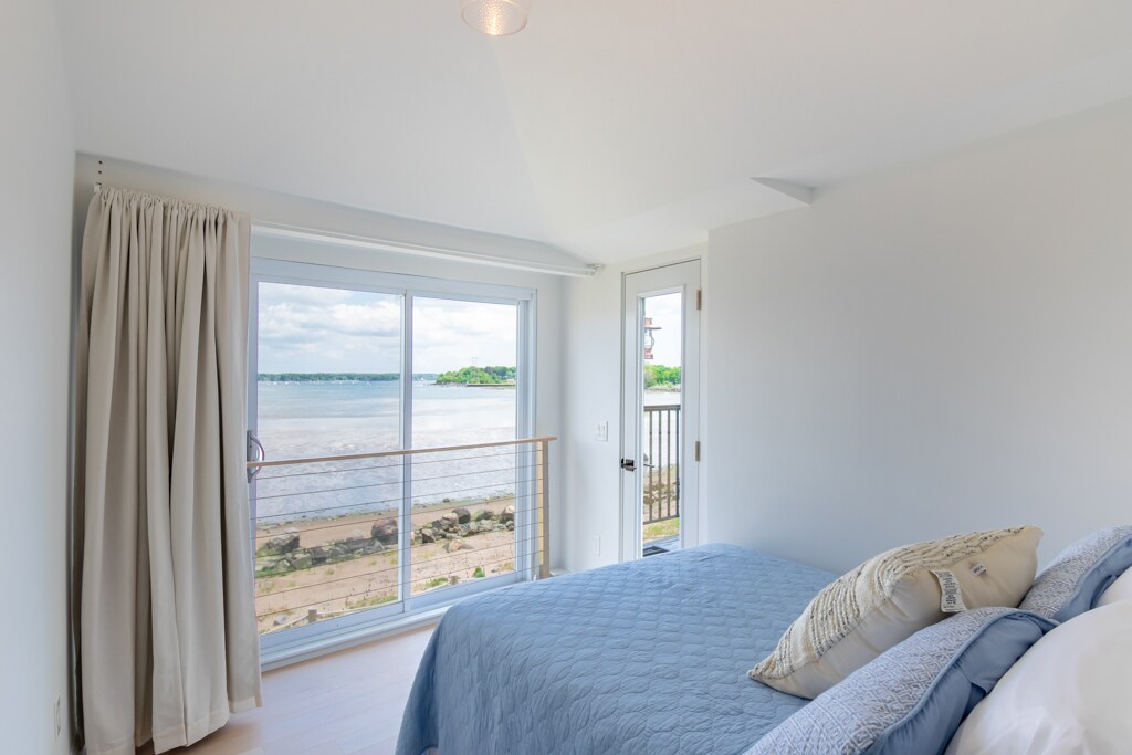 A serene bedroom features a spacious bed with a blue coverlet. Large windows provide expansive views of the water, with natural light illuminating the room. A door leads to an outdoor space, complementing the tranquil ambiance.