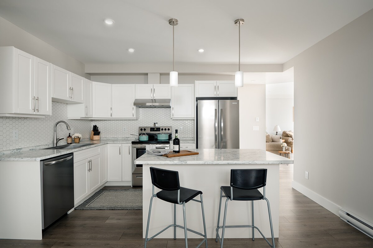 A modern kitchen features white cabinetry complemented by a gray countertop. Stainless steel appliances include a refrigerator and oven. Two black stools are positioned at the kitchen island, which is illuminated by pendant lights, creating a bright and functional cooking space.