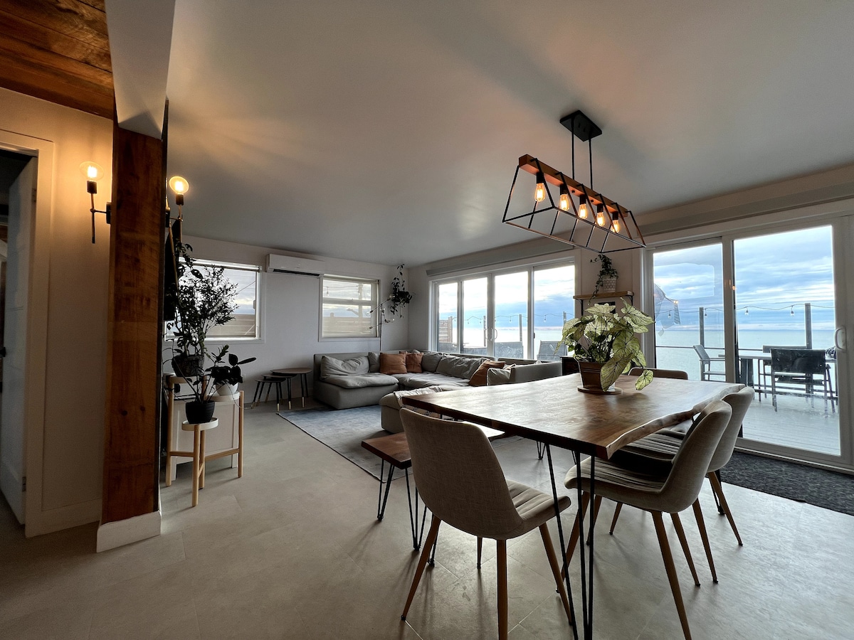 A spacious living area is presented with large windows that frame views of the river. Comfortable seating arrangements, including a sectional sofa, create a welcoming environment. A dining table with six chairs is centrally positioned, illuminated by a modern light fixture overhead.