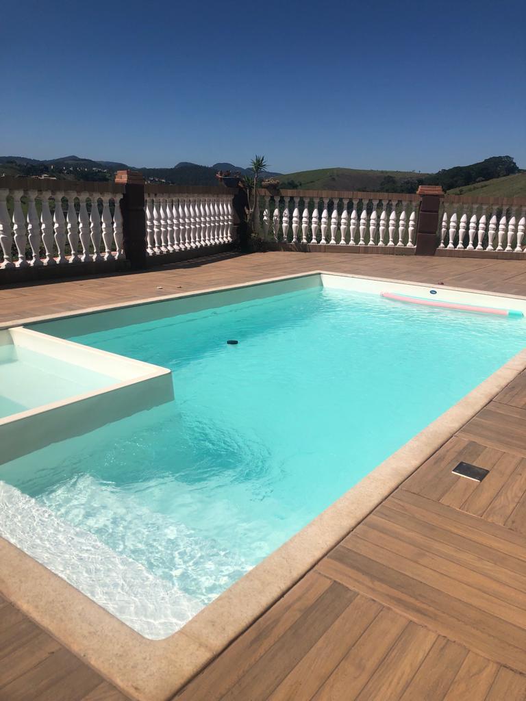 A tranquil pool occupies a spacious wooden deck, featuring clear turquoise water that reflects the bright blue sky above. The surrounding railing provides a panoramic view of the distant hills, enhancing the serene atmosphere of the outdoor space.