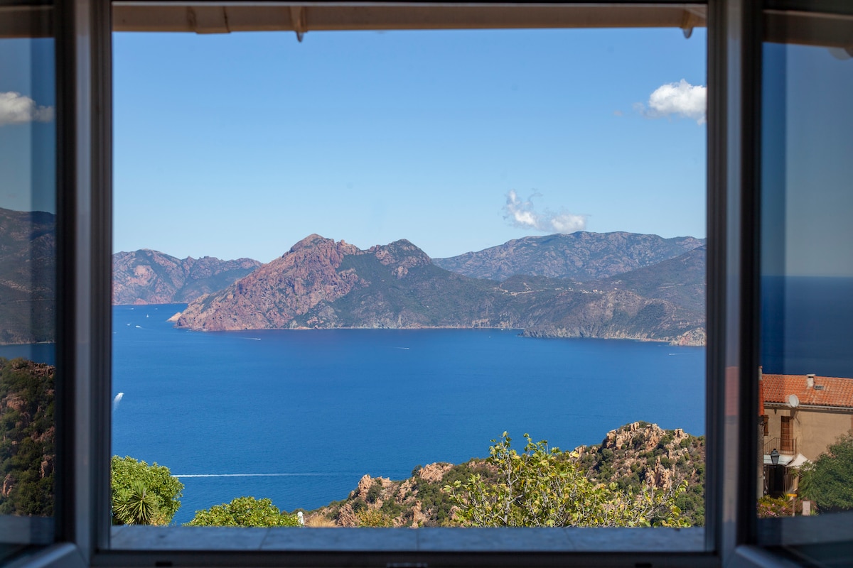 A panoramic view is presented through a large window, showcasing the blue waters of the Gulf of Porto and the surrounding hills. The landscape includes rocky mountains contrasting with a clear sky, enhancing the serene coastal scenery.