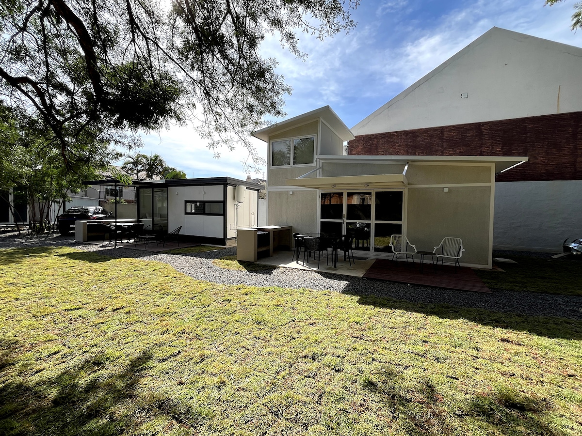 A spacious outdoor area is displayed, featuring a covered patio with a barbecue grill and several chairs. Lush green grass surrounds the seating area, and nearby structures are visible in the background, providing a sense of privacy and comfort.